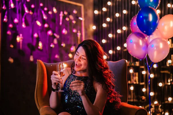 Bonita jovencita delante de la cámara en el sofá disfrutando de la fiesta de cumpleaños sosteniendo una copa de champán y chispa —  Fotos de Stock