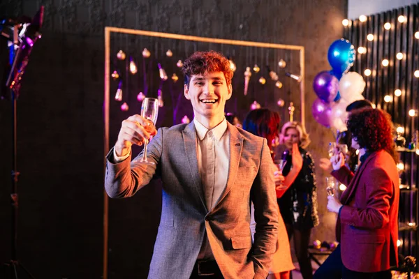 Attraente e divertente ragazzo con i capelli ricci che tiene un bicchiere di champagne davanti alla telecamera alla grande festa si gode il momento Foto Stock