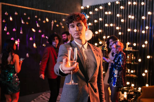Attraente e divertente ragazzo con i capelli ricci che tiene un bicchiere di champagne davanti alla telecamera alla grande festa si gode il momento Fotografia Stock