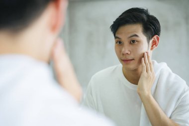 Asyalı genç adam banyo aynasında yüz kremiyle sivilceleri temizledikten sonra yüzlerine dokunuyor..