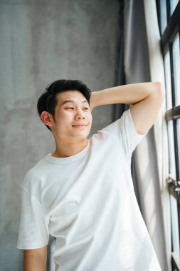 Yataktan kalktıktan sonra kollarını açıp pencereden dışarı bakan Asyalı genç adam..