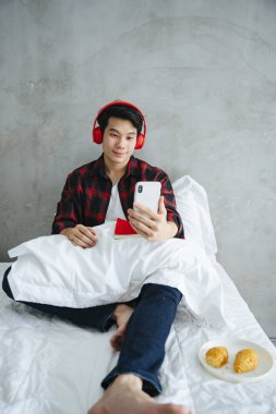 Asyalı genç adam kablosuz kulaklıkla ve yatakta akıllı fonla müzik dinliyor..