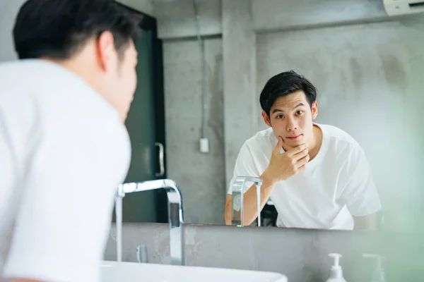 Asian young man cleaning face at faucet in bathroom and looking in mirror.