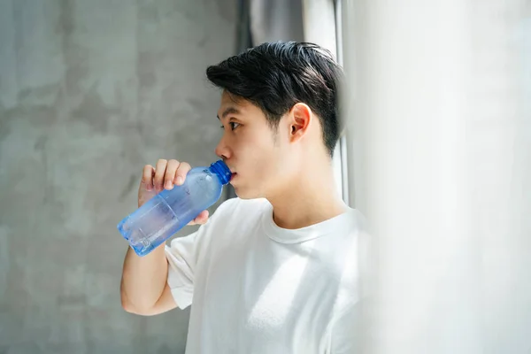 Asian young man in white shirt drinking from bottle of water at home.