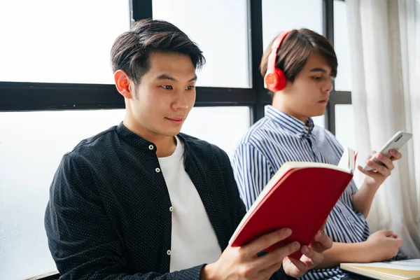 Couple of asian LGBTQ+ men spending time reading a book and listing music with wireless headphone and smartphone.