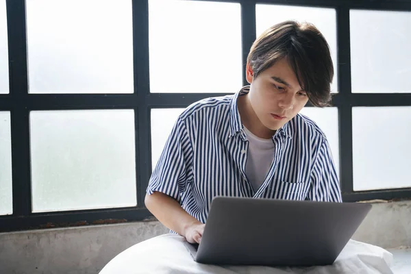 Asian young man in casual looking working on laptop computer near window at home