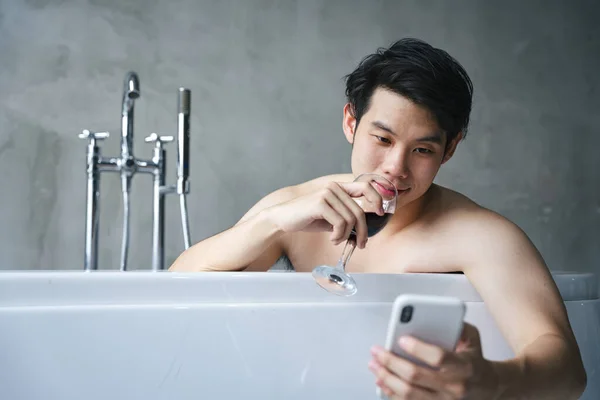 浴槽でアジアの若い男ワインを飲み スマートフォンを使用して — ストック写真