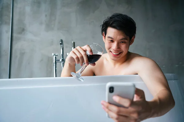 Asian young man in bathtub drinking wine and using smartphone.
