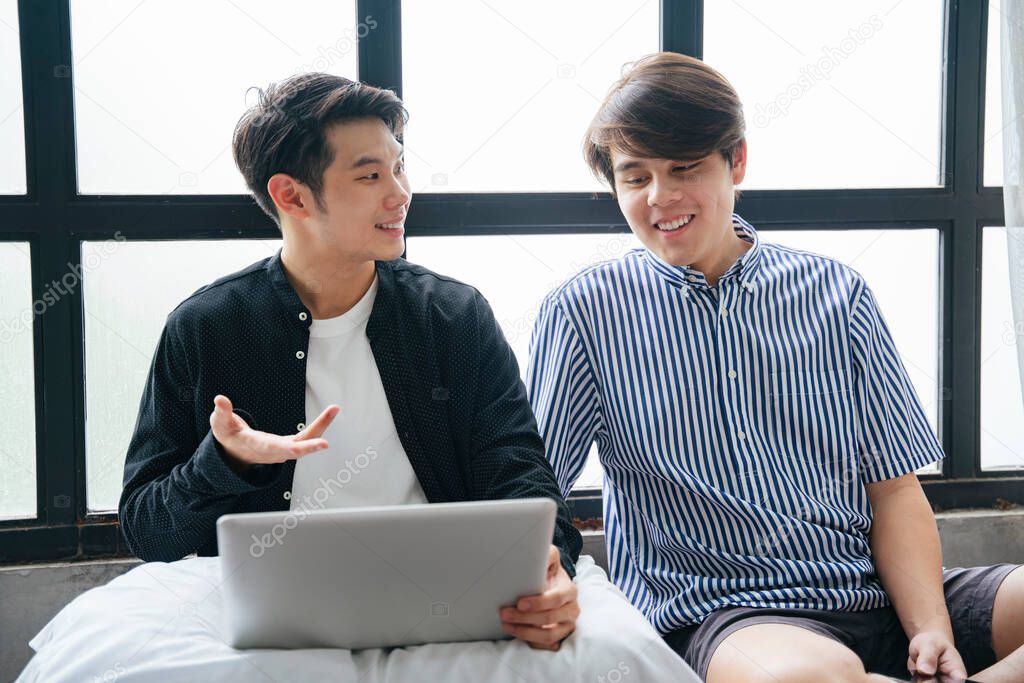 Couple of asian LGBTQ+ men sit together and talking while using laptop.