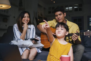 Oturma odasındaki kanepede oturup birlikte eğlenen mutlu bir aile. Anne gülüyor, baba gitar çalıyor, çocuk komik bir şey yapıyor..