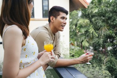 Asyalı bir adam, balkonda birlikte şarap içerken kız arkadaşıyla konuşuyor..