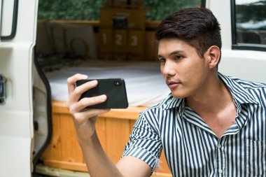 Asyalı bir adam kamp alanında CV arabasının yanında akıllı telefonuyla selfie çekiyor..