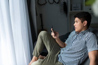 Asyalı adam evdeki dijital akıllı telefonu kullanarak kanepede oturuyor..