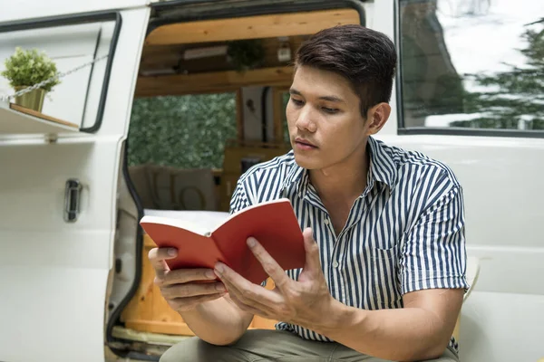 Asian man reading a book near CV car at camping site outdoors.