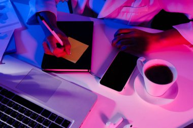 Üst Manzara - iş kadını dizüstü bilgisayarı, kitabı, kahvesi, telefonu neon renkli ışıkta çalışıyor.