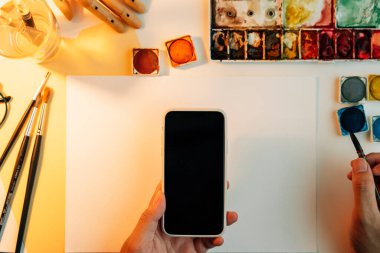 Üst görünüm - referans bulmak için renkli boya ekipmanları ve akıllı telefon ile çalışan sanatçı masası.