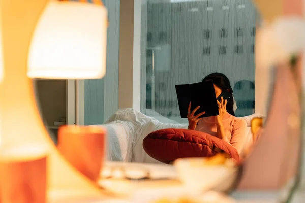 Woman reading a book before sleep in bedroom with lamp light on.