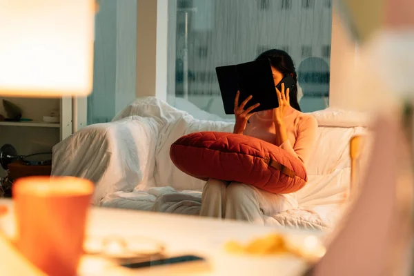 Woman reading a book before sleep in bedroom with lamp light on.