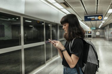 Asyalı Taylandlı sırt çantalı kadın metro istasyonunda tren beklerken akıllı telefon kullanıyor..