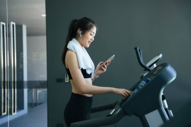 Sağlıklı Asyalı kadın koşu bandında çalışırken akıllı telefondan şarkı seçiyor..