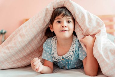 Heyecanlı küçük kız yatak odasında battaniyenin altında komik surat yapıyor..