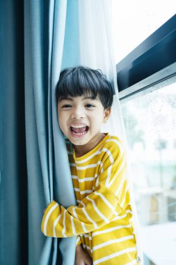 Pencerenin yanındaki mavi perdenin arkasında beyaz kazaklı neşeli çocuğun portresi..