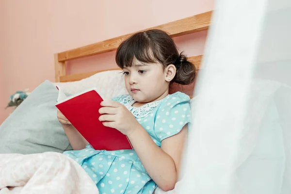 Adorable girl in blue dress read a book before bed at bedroom.