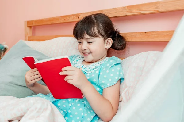 Adorable girl in blue dress read a book before bed at bedroom.