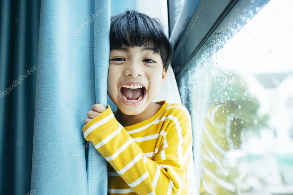 Portrait of cheerful funny boy in white sweater behind blue curtain near window.