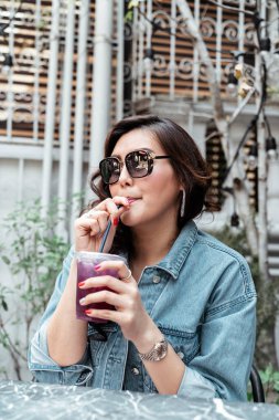 Kot pantolon giyen Asyalı Taylandlı kadın güneş gözlüğü takıyor kafede samanlı plastik bardaktan üzüm suyu içiyor..