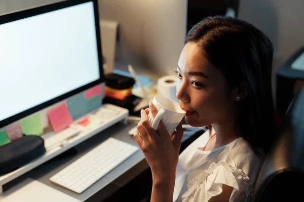 Asian thai woman drinking and sipping hot coffee from a cup, sitting infront of computer at office. Night shift time.