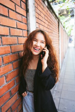 Tuğla duvarın yanında konuşan siyah ceketli genç Asyalı kadının moda portresi..