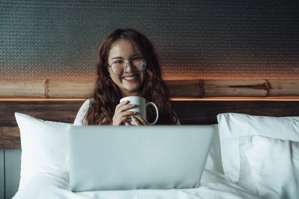 Asian woman enjoy hot coffee for breakfast and using laptop computer on bed.