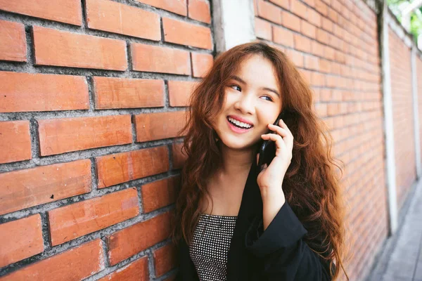 レンガの壁の近くの電話で話して黒のジャケットを身に着けている若いアジアの女性のファッション肖像画 — ストック写真
