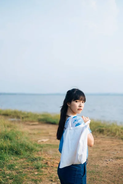 穿着白蓝相间的领带的黑发女郎背着帆布袋走在海边的草地上 转身对着相机拍了照 — 图库照片