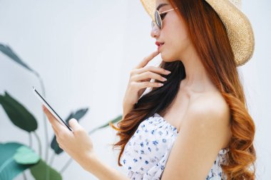 Yan görünüş - Asyalı güzel gezgin kadın güneş gözlüğü ve şapka takıyor akıllı telefon kullanıyor..