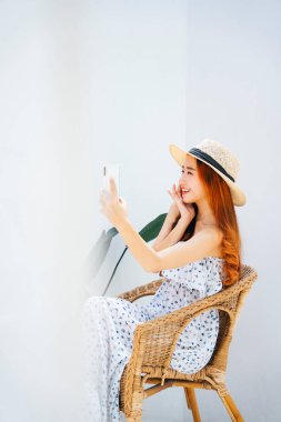 Yan görüş - Asyalı güzel gezgin kadın sandalyede akıllı telefon kullanarak şapka takıyor..