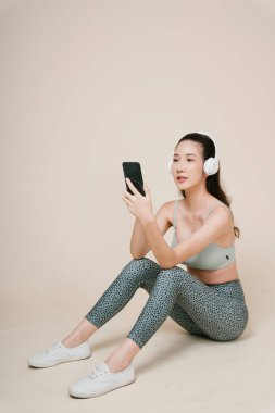 Yerde oturan Asyalı sporcu kadının portresi kulaklık ve akıllı telefonla müzik dinliyor..