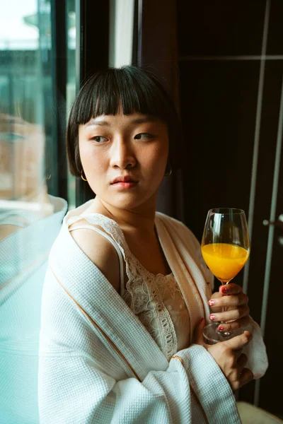 Asian short hair woman in white bathrobe enjoy breakfast with orange juice in hotel room.