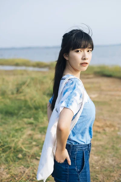 穿着白蓝相间领带的黑发姑娘站在海边的草地上 提着帆布袋 摸着她的背口袋 — 图库照片