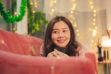 Genç Asyalı kadın yılbaşı süslemeli bir odada koltuğa uzanmış akıllı telefon kullanıyor..