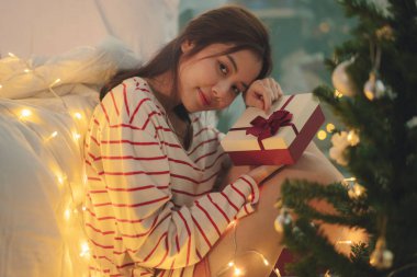 Güzel, genç Asyalı kadın. Siyah saçlı. Yatak odasında Noel ışığı ve hediye kutusu var..