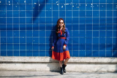 Mavi ve kırmızı retro elbiseli ve bereli esmer kadın mavi duvarda güneş ışığında duruyor..