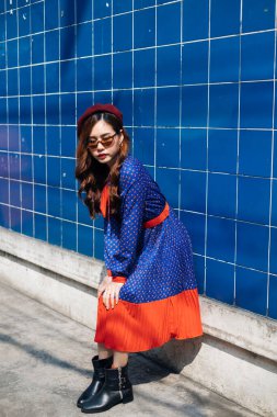 Dalgalı esmer kadın, mavi ve kırmızı retro elbiseli ve bereli güneş gözlüklü mavi duvarda duruyor..