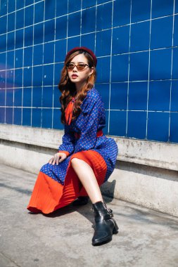 Mavi ve kırmızı retro elbisesi ve beresi olan dalgalı esmer kadın güneş ışığında seksi duruşuyla mavi duvarda oturuyor..