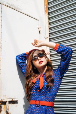 Kırpılmış mavi ve kırmızı retro elbiseli esmer kadın görüntüsü ve güneş gözlüğü eski depodaki metal kepenkli süpermodel gibi duruyor..