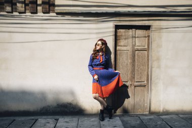 Dalgalı esmer, mavi ve kırmızı retro elbiseli kadın yokuşun üstünde duruyor ahşap kakanın önünde..