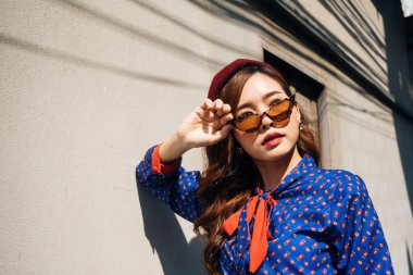 Mavi ve kırmızı retro elbiseli, dalgalı esmer kadının orta boy portresi güneş ışığında eski evin önünde duruyor..