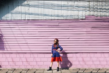 Mavi ve kırmızı retro elbiseli dalgalı esmer kadın. Pembe tuğla ve güneş ışığında tahta duvarın önünde duruyor. Manken gibi davran..