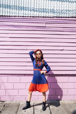 Mavi ve kırmızı retro elbiseli dalgalı esmer kadın. Pembe tuğla ve ahşap duvarın önünde duruyor. İçi de güneş ışığında süpermodel..